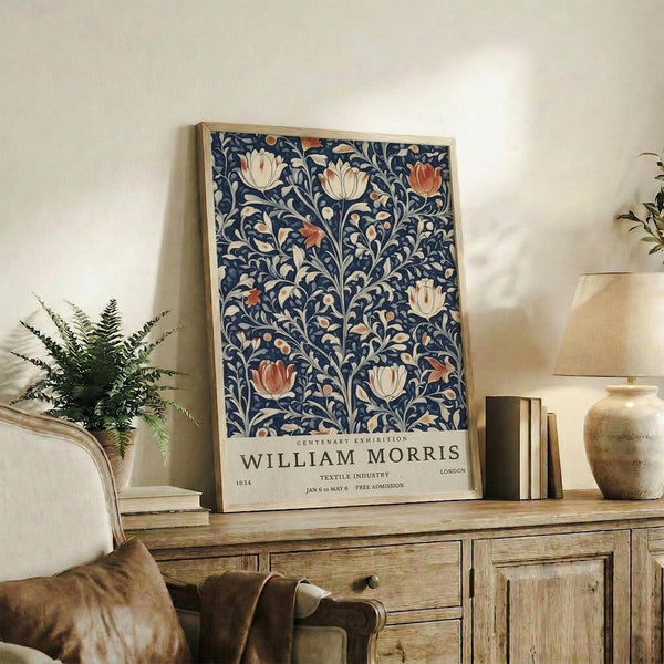 May include: Framed William Morris exhibition poster with intricate navy blue floral pattern featuring cream and coral tulips and botanical motifs, displayed on rustic wooden sideboard alongside fern plant, ceramic vases, and books in warm-lit living room with beige armchair and neutral walls