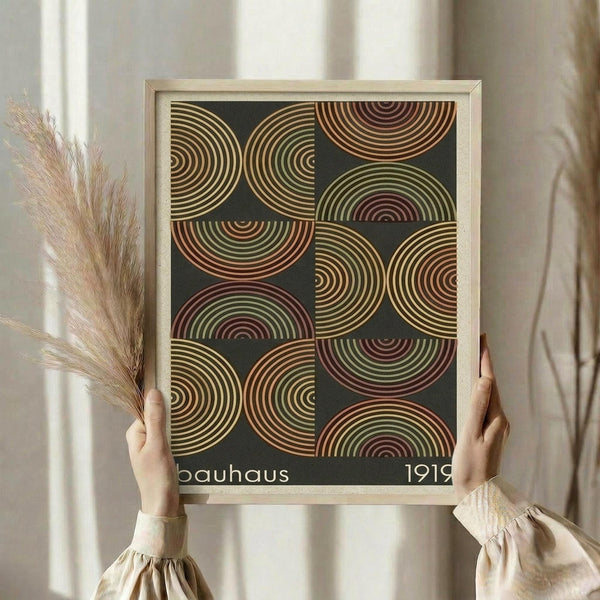 May include: Person holding framed geometric poster featuring overlapping semicircular patterns in warm earth tones of orange, green, and burgundy on dark background. Light wood frame, cream matting, displayed against soft neutral curtains with dried pampas grass visible to the side.