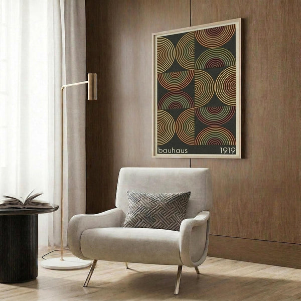 May include: Framed geometric poster with colorful concentric circles in earth tones on dark background, displayed on wood-paneled wall above modern cream armchair with patterned pillow. Room features brass floor lamp, side table with open book, and neutral contemporary decor.
