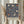 May include: Hands holding a framed vintage exhibition poster featuring William Morris floral textile design with navy blue background and cream, coral, and rust colored tulips and flowing botanical motifs. Light wooden frame against soft neutral curtains with dried wheat stems visible on left side.