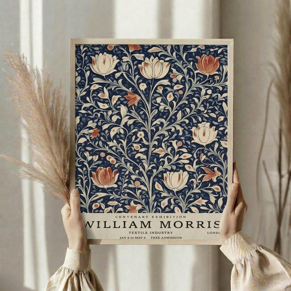 May include: Hands holding a framed vintage exhibition poster featuring William Morris floral textile design with navy blue background and cream, coral, and rust colored tulips and flowing botanical motifs. Light wooden frame against soft neutral curtains with dried wheat stems visible on left side.