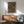 May include: Framed geometric poster with colorful concentric circles in red, green, and yellow on dark background, displayed above wooden sideboard with table lamp in modern living room setting. Mid-century style chair with sage green upholstery visible in foreground. Natural sunlight creates shadows on white wall.