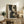 May include: Abstract geometric artwork with dark green and beige shapes displayed in light wood frame on rustic wooden sideboard. Room scene shows cream upholstered chair, potted fern, ceramic vases, scattered leaves, and neutral wall. Artwork features curved and angular forms in earthy tones with textured, weathered appearance.