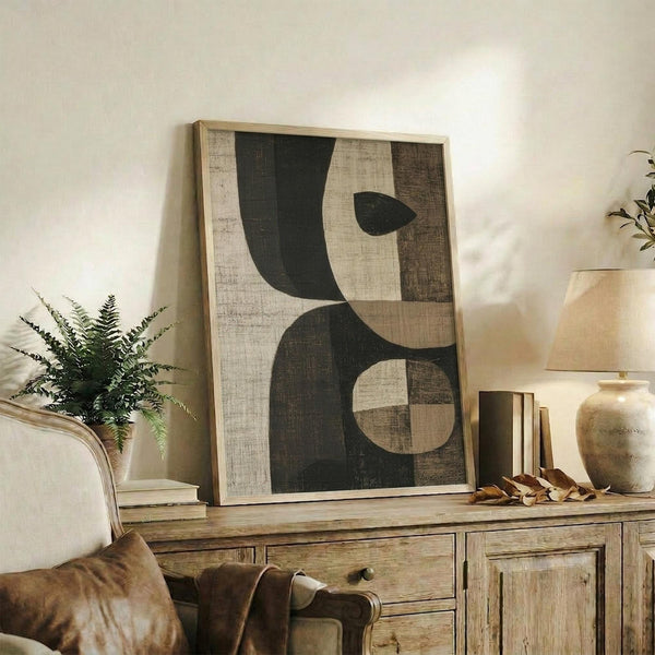 May include: Abstract geometric artwork with dark green and beige shapes displayed in light wood frame on rustic wooden sideboard. Room scene shows cream upholstered chair, potted fern, ceramic vases, scattered leaves, and neutral wall. Artwork features curved and angular forms in earthy tones with textured, weathered appearance.
