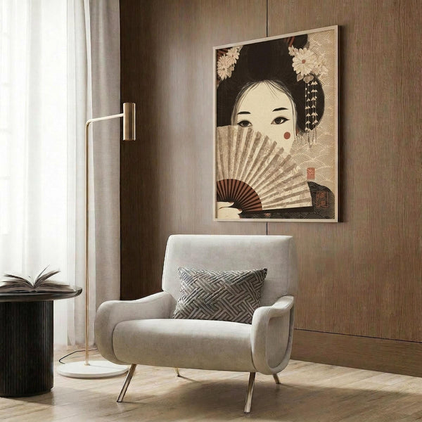 May include: Japanese-inspired artwork showing a woman with dark hair adorned with flowers, holding an ornate fan that partially covers her face. The poster features warm beige and brown tones with decorative wave patterns. Displayed in a light wood frame on wood-paneled wall above a cream upholstered armchair with geometric patterned pillow, beside a brass floor lamp and dark round side table with open book.