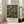 May include: Framed botanical poster with orange fruit tree design on deep blue background, displayed on wooden floor next to beige upholstered chair in bright room with white walls and windows. The vintage-style artwork features detailed branches, green leaves, and round oranges in warm tones, with exhibition text below the illustration.