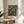 May include: Framed vintage exhibition poster featuring orange fruit tree with green foliage against teal background, displayed on round wooden side table next to beige upholstered chair. Natural sunlight streams across white walls, with plant leaves visible in corner. Poster shows botanical illustration in Arts and Crafts style with exhibition text below artwork.