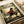 May include: Framed anime poster print displayed on white fabric surface with rolled paper tube nearby. Features dark-haired samurai character in red and black robes holding katana sword, set against bamboo forest background with dappled sunlight. Japanese text at top, English subtitle below. Warm earth tones with green foliage details.