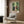 May include: Framed anime poster displaying three characters walking through a bamboo forest - one in dark blue robes, one in pink kimono, and one in red jacket. The artwork features lush green bamboo with Japanese text below. Poster hangs on wood-paneled wall above a beige upholstered armchair with geometric throw pillow. Modern floor lamp and side table with open book complete the cozy reading nook setting.