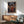 May include: Framed anime poster featuring a dark-haired samurai character in red robes holding a sword, set against a traditional Japanese street scene with lanterns and crowds at dusk. The poster hangs above a wooden sideboard with a white ceramic table lamp, next to a mid-century modern teal upholstered armchair with wooden frame. Warm sunlight streams across white walls creating geometric shadows in this minimalist living room setting.
