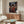 May include: Framed anime poster mounted on white wall above wooden credenza in modern living room. Poster shows dark-haired samurai character in red kimono holding sword on traditional Japanese street at dusk with lanterns and crowds. Room features white curved chair, oval glass coffee table, brass floor lamp, and neutral decor with natural lighting.