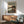 May include: Framed anime poster mounted on white wall above wooden credenza with lamp and mid-century modern teal chair. Poster features samurai character with sword in earth-toned illustration style with Japanese and English text. Warm natural lighting creates shadows across minimalist living room setting with hardwood floors.