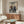 May include: Framed anime poster on white wall above wooden sideboard in modern living room. Poster shows two samurai warriors dueling over stylized ocean wave in vintage Japanese art style with blue, cream and red colors. White curved chair and glass coffee table visible in foreground with warm natural lighting.