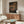 May include: Anime poster print displayed above wooden sideboard in modern living room setting. Three animated characters sit at table in traditional Japanese tea house scene with warm lighting and architectural details. Poster has thin frame, surrounded by white chair, floor lamp, and glass coffee table reflecting blue and yellow tones.