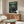 May include: Framed anime poster displayed above wooden sideboard in modern living room with cream chair and glass coffee table. Poster shows three samurai characters in bamboo forest setting with green and earth tones. White walls, natural lighting, mid-century modern furniture styling.