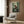 May include: Framed anime poster featuring a dark-haired samurai character in red and brown clothing wielding a sword, leaping through a green bamboo forest. The poster hangs on a wood-paneled wall above a beige upholstered armchair with geometric patterned pillow. Modern living room setting includes a brass floor lamp and dark round side table with open book.