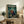 May include: Framed anime poster displayed on rustic wooden sideboard in cozy living room setting. Artwork shows three samurai characters in traditional clothing wielding swords in bamboo forest scene with teal and green tones. White text at top reads "SAMURAI CHAMPLOO" with Japanese characters below. Poster leans against white wall beside fern plant, ceramic vases, and stacked books on weathered wood furniture.