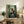 May include: Framed anime poster displayed on rustic wooden sideboard in cozy living room setting. Shows samurai character in red clothing wielding sword among green bamboo forest background with Japanese text. Natural wood frame, surrounded by fern plant, ceramic vase, books, and beige upholstered chair with brown throw pillow.