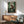 May include: Framed anime poster showing a samurai character with dark hair in red jacket and dark pants, wielding a sword among green bamboo forest. Japanese text at top and bottom. Wooden frame hangs above mid-century modern furniture including teal upholstered chair with wooden arms, wooden sideboard, and white table lamp. Sunlight creates shadows on white wall.