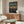 May include: Japanese anime-style poster featuring silhouetted swordsman standing on traditional rooftops at sunset, displayed in thin wooden frame above mid-century walnut credenza. Room shows modern minimalist decor with cream curved chair, oval glass coffee table, white table lamp, and neutral walls with natural lighting.