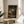 May include: Framed anime poster displayed on round wooden side table next to beige upholstered chair. Artwork shows character in red jacket sitting in traditional Japanese room with tatami mats, wooden architecture, and calligraphy scrolls. Japanese and English text at bottom reads "Samurai Champloo" and "A Hustle to be Free." Natural lighting creates soft shadows on white wall behind. Plant leaves visible in corner.