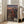 May include: Framed anime poster leaning against white wall next to beige sofa in bright living room. Shows three characters on rooftop overlooking traditional Japanese town at sunset - woman in red kimono, tall man in dark blue robes, and seated figure in burgundy clothing. Warm orange and purple sky with detailed cityscape below. Light wood frame, approximately 2-3 feet tall, positioned on hardwood floor near large windows.