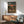 May include: Framed anime poster hanging on white wall above wooden sideboard, showing three characters walking on dirt path toward sunset with mountains in background. Japanese text at top. Mid-century modern room with mint green upholstered chair, white table lamp, and natural lighting creating shadows on wall.