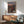 May include: Framed anime poster showing three characters on rooftop at sunset with traditional Japanese town below, displayed above wooden sideboard with white table lamp in modern living room with teal upholstered chair and round coffee table, natural sunlight casting shadows on white wall