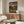 May include: Framed anime poster hanging above wooden sideboard in modern living room. Poster shows three silhouetted figures walking on dirt path toward sunset with Japanese text. Room features white curved chair, glass-top coffee table, white table lamp, and neutral walls with natural lighting.
