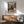 May include: Framed anime poster hanging on white wall above wooden sideboard in modern living room. Features samurai character with long dark hair in red and blue clothing wielding sword in mountainous landscape. Warm sunlight streams across mint green mid-century chair and cream table lamp. Natural wood frame complements furniture's warm tones.
