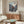 May include: Framed anime poster displayed above wooden sideboard in modern living room setting. Poster features samurai character in red garment wielding sword amid dynamic action scene with Japanese text. Room includes white curved chair, oval glass coffee table, floor lamp, and neutral beige walls with natural lighting.