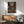May include: Framed anime poster showing two samurai warriors with swords in traditional Japanese street setting with crowd watching, displayed above wooden sideboard with lamp in modern living room with mint green armchair and natural sunlight streaming across white wall.