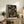 May include: Framed anime poster displayed on rustic wooden sideboard in cozy living room setting. Artwork shows samurai character with black hair in red jacket running through traditional Japanese street with glowing paper lanterns. Poster features Japanese text and evening alley scene. Natural wood frame, surrounded by potted fern, ceramic vase, stacked books, and beige upholstered furniture with warm lighting.