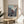 May include: Framed anime poster displayed on round wooden side table next to beige upholstered chair. Poster shows dynamic samurai character in red jacket wielding sword amid action scene with traditional Japanese buildings and crowd. Japanese text at top and bottom. Natural lighting creates soft shadows across neutral-toned room setting with plant visible in corner.