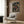 May include: Framed anime poster mounted on wood-paneled wall above beige upholstered armchair with geometric pillow. Poster shows samurai character in red jacket walking through lantern-lit Japanese street at night. Modern reading nook with gold floor lamp and dark side table with open book. Warm lighting creates cozy atmosphere in contemporary interior space.