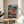 May include: Framed One Piece anime poster featuring Luffy in his signature straw hat and red vest, fist glowing with power against a stormy ship background. Japanese text visible below character. Natural wood frame displayed on round wooden side table next to beige upholstered chair, with plant leaves and soft window lighting creating shadows in minimalist room setting.