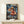 May include: Framed One Piece anime poster displayed on light wood shelf against cream wall. The artwork shows Luffy in his signature straw hat and red vest with flaming fist, set against stormy ship scene with lightning. Japanese and English text below character. Small white gift box sits beside the wooden-framed print on minimalist shelf.