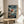 May include: Framed One Piece anime poster displayed on wooden round side table next to beige upholstered chair. Poster shows Luffy in red shirt and straw hat leaping from small wooden boat across stormy ocean with dramatic cloudy sky. Japanese text and "Journey Beyond the Horizon" subtitle visible. Natural lighting creates soft shadows across neutral-toned room setting with plant leaves visible in corner.