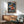 May include: Framed One Piece anime poster featuring Luffy in red vest and straw hat with flaming fist on stormy ship deck background, displayed above wooden sideboard with white ceramic lamp in modern living room with teal upholstered chair and natural lighting casting shadows on white wall.