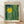 May include: Framed poster print leaning against white wall on light wood floor. The artwork features a bright yellow sun surrounded by stylized forest with vertical tree trunks in teal, green and yellow tones, decorated with dotted patterns and organic shapes. The print has "YAYOI KUSAMA" at top, "Modern Art" and "1964" at bottom in black text on cream background. Natural wood frame visible in warm lighting with soft shadows.