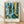 May include: Framed art print leaning against white textured wall on light wood floor. Abstract artwork features layered blue and teal tones with geometric patterns, organic leaf shapes, golden yellow accents, and dark contrasting elements. Mixed media style with grid patterns, dots, and flowing forms suggesting underwater or tropical themes. Natural wood frame with white matting displaying artist attribution text below the image.