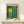 May include: Framed art print with yellow sun and green forest design in modern abstract style, displayed on light wood shelf next to small wrapped gift with feather accent against white wall