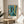 May include: A framed reproduction of Paul Klee's abstract artwork featuring vibrant blues, teals, and golden yellows with overlapping geometric patterns, botanical motifs, and textured brushwork. The print sits on a round wooden side table next to a beige upholstered chair, with plant shadows cast across white walls and natural sunlight streaming through nearby windows.