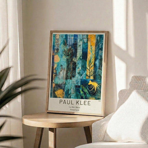 May include: A framed reproduction of Paul Klee's abstract artwork featuring vibrant blues, teals, and golden yellows with overlapping geometric patterns, botanical motifs, and textured brushwork. The print sits on a round wooden side table next to a beige upholstered chair, with plant shadows cast across white walls and natural sunlight streaming through nearby windows.
