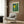 May include: Cream-colored poster with yellow sun and green bamboo forest design hanging on wood-paneled wall above beige upholstered armchair with geometric pillow. Modern brass floor lamp and dark round side table with open book complete the neutral-toned reading nook setting.