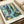May include: Framed art print featuring Paul Klee's abstract composition with tropical foliage, geometric patterns, and layered textures in blues, teals, yellows and oranges. The print shows palm fronds, grid patterns, and collage-like elements typical of Klee's style. Displayed in a light wood frame on cream colored paper, sitting on white bedding with soft natural lighting.