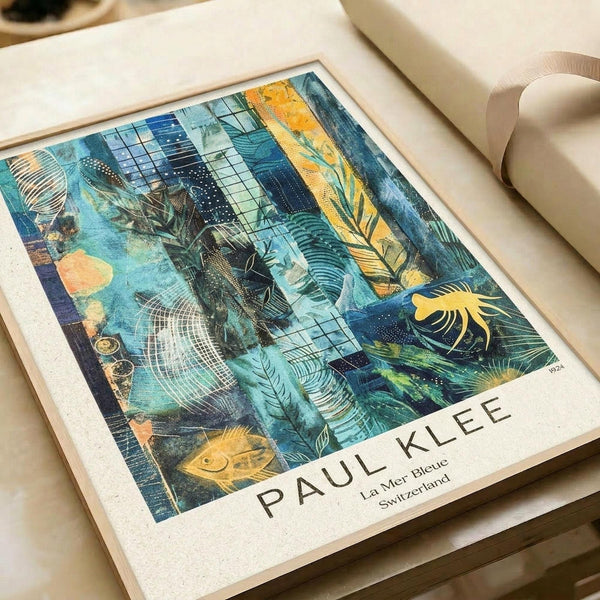 May include: Framed art print featuring Paul Klee's abstract composition with tropical foliage, geometric patterns, and layered textures in blues, teals, yellows and oranges. The print shows palm fronds, grid patterns, and collage-like elements typical of Klee's style. Displayed in a light wood frame on cream colored paper, sitting on white bedding with soft natural lighting.