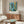 May include: A framed art print featuring an abstract tropical composition with blue, teal, yellow and green botanical elements including palm fronds and geometric patterns. The poster hangs above a mid-century modern walnut credenza with brass hardware. A white curved chair sits beside an oval glass-top coffee table with wooden base, and a brass floor lamp with white shade stands nearby. The room has cream walls and natural lighting.