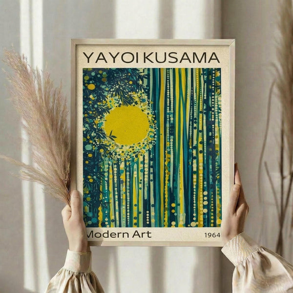 May include: Person holding framed poster print in light wood frame against white curtained window. Artwork features bright yellow sun with dark blue and teal vertical streaks, dots, and botanical elements. Person wears cream colored outfit with puffy sleeves, holds dried pampas grass in left hand. Natural lighting creates soft shadows on white wall background.