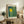 May include: Framed art print with yellow sun and vertical blue-green streaks in polka dot pattern, displayed on rustic wooden sideboard beside fern plant, books, and ceramic vases in bright living room setting with beige sofa and white walls